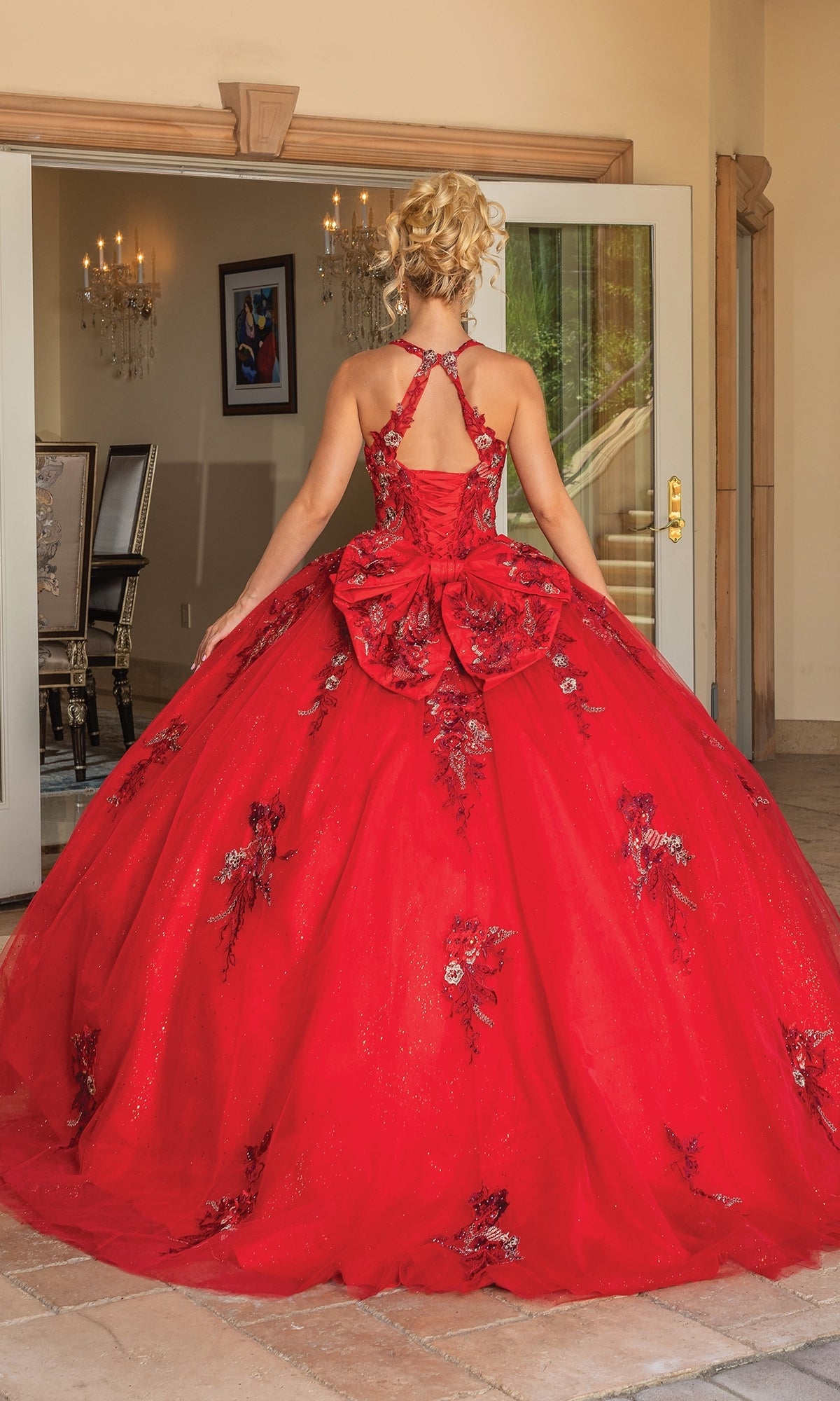 A woman wears the Quinceanera Dress 1761 By Dancing Queen, a formal red gown with floral embellishments and a large back bow, as she stands facing open double doors.